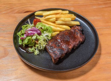 Smoky BBQ domuz pirzolası, patates kızartması ve taze salata, siyah tabakta, ahşap masa kenarında Amerikan konforlu yemekleri.