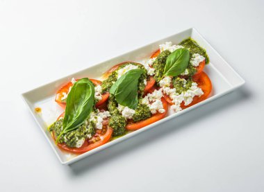 Dilimlenmiş domatesli Caprese Salatası, Cottage Cheese, Pesto ve taze fesleğen beyaz dikdörtgen tabakta beyaz arka plan görünümünde izole edilmiş.