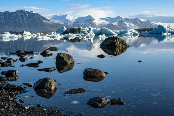 Black rocks and boulders reflected in the water of Jokulsarlon glacier lagoon, icebergs and Fellsfjall mountains in the background, Iceland, Vatnajokull National Park, near Route 1 / Ring Road