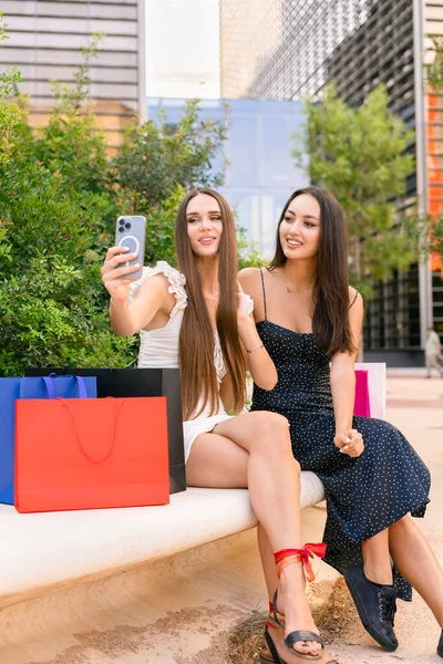드레스를 벤치에 쇼핑백으로 셀카를 스마트폰을 사용하는 매력적 아가씨들 — 스톡 사진