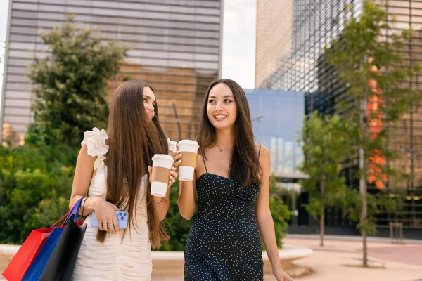 Positive female friends with paper bags and coffee to go walking in city after successful shopping on weekend
