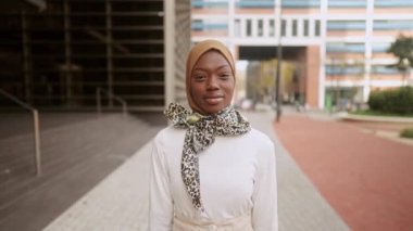 Görüntüyü yakınlaştırın ve şık gündelik kıyafetlerle şık başörtüsü ve eşarbı ile siyah bir kadın görüntüsünü uzaklaştırın. Kamera karşısında başını çevirip gülümserken aynı zamanda şehir caddesindeki modern binanın yanında duruyor.