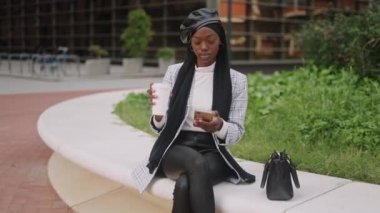 Bere ve başörtüsü takmış, cep telefonuyla sosyal medyada gezinen ve parkta otururken kahve yudumlayan Müslüman kadınlar.