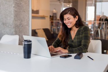 Ofiste çalışırken bir fincan sıcak içecek ve dizüstü bilgisayarla masada oturan pozitif Asyalı kadın işçi.