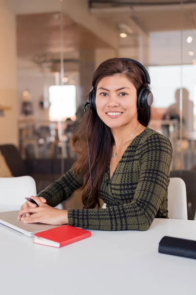 Young Asian woman in headset sitting at table with netbook and notepad looking at camera in office