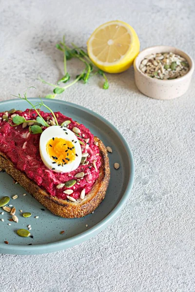 Whole grain toast with beetroot hummus, seeds, egg and microgreens (sprouted pea sprouts) on a light gray background. The concept of healthy eating..