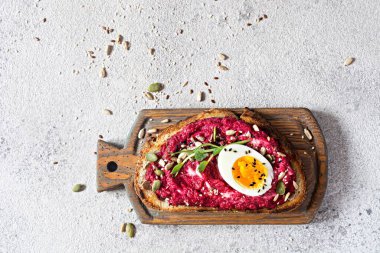 Açık gri bir zemin üzerinde pancar kökü humus, tohumlar, yumurta ve mikro yeşillikler (bezelye filizi filizleri) ile tam tahıllı tost. Sağlıklı beslenme kavramı. Yaşasın 2023 'ün mor rengi.