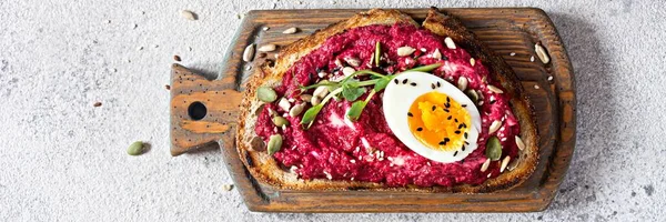 Whole grain toast with beetroot hummus, seeds, egg and microgreens (sprouted pea sprouts) on wooden board on a light gray background. The concept of healthy eating. Viva magenta color of the year 2023