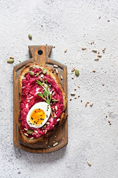 Whole grain toast with beetroot hummus, seeds, egg and microgreens (sprouted pea sprouts) on wooden board on a light gray background. The concept of healthy eating. Viva magenta color of the year 2023