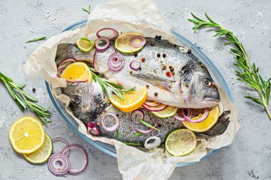 Fresh fish dorado. Raw dorado with herbs, spices and lemon and lime slices ready to cook on a gray background. Top view.  Fresh fish in a glass baking dish  with salt, herbs and pepper.