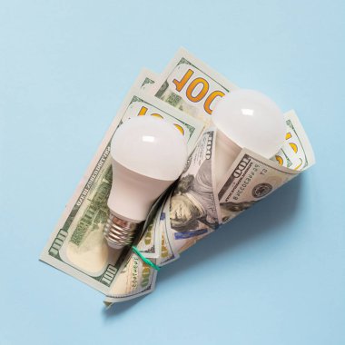 Mavi arka planda Amerikan para birimiyle (dolar) enerji tasarrufu ampulleri. Elektrik için ödeme, enerji pazarının mali durumu, enerji fiyatlarındaki artış.