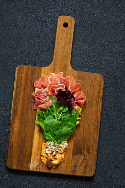 stock image Salad of jamon (prosciutto) spinach and lettuce with crispy savory bread, nuts, microgreens on black background. Flat lay of food in form of light bulb on board. Food energy concept, top view.