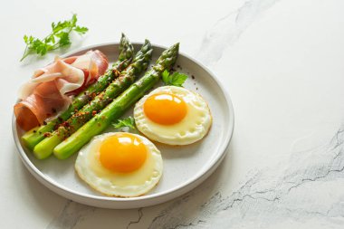 Taze kuşkonmaz ve jambon (jambon) ile kızarmış kahvaltı yumurtaları, beyaz arka planda bir tabakta otlar. Nefis bir kahvaltı..