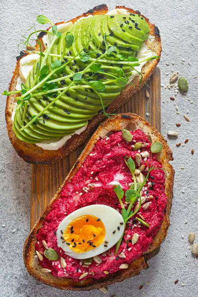 Whole grain toast with cream cheese, avocado, beetroot hummus, seeds, egg and microgreens (sprouted pea sprouts) on a light gray background. Healthy food concept.