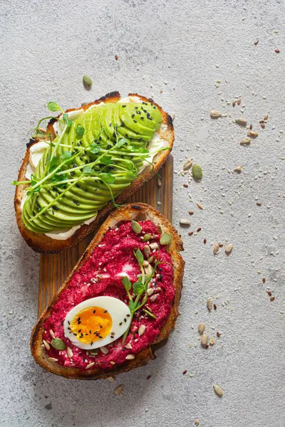 Whole grain toast with cream cheese, avocado, beetroot hummus, seeds, egg and microgreens (sprouted pea sprouts) on a light gray background. Healthy food concept.