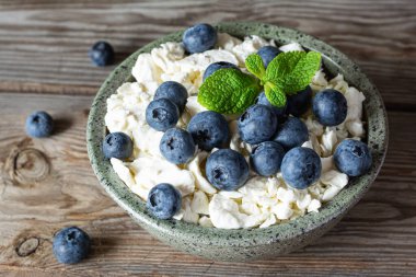 Bir kase süzme peynir, yoğurt, taze böğürtlen (yaban mersini) ve ahşap zemin üzerinde taze nane. Lezzetli kahvaltı, sağlıklı yemek..