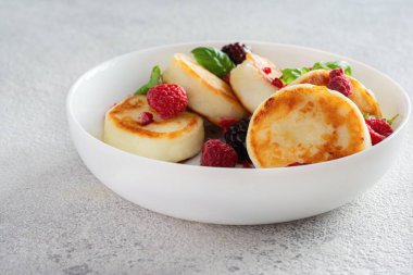 Taze süzme peynir ya da ricotta krepleri (şurup, börek) mavi arka planda taze böğürtlenli beyaz bir tabakta. Lezzetli sağlıklı kahvaltı. Ukrayna mutfağı, tatlı yemekler.