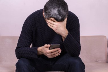 Orta yaşlı bir adam telefonda üzücü haberler alıyor. İnternette haber okuyan endişeli bir adam, telefon ekranına bakıyor, beyaz tenli, evdeki kanepede oturuyor, ciddi ve üzgün.