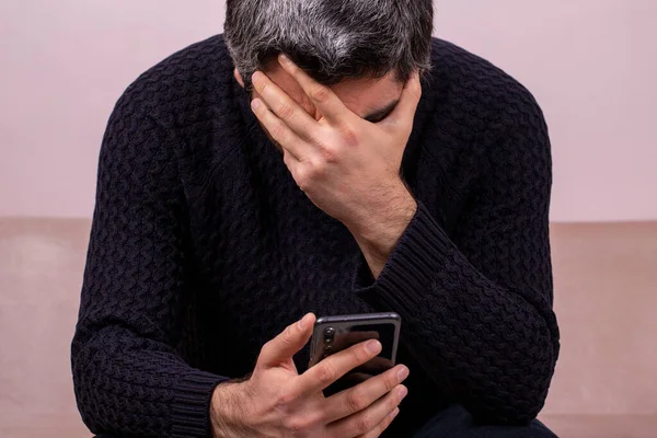 Middle-aged man receiving sad news on the phone. Concerned man reading news online looking at phone screen, Caucasian sitting on sofa at home, serious and sad