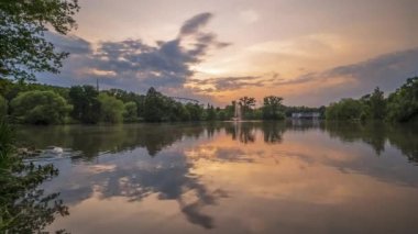 Gün batımında hareket eden bulutları, yüzen ve göldeki bulutları yansıtan bir kuğu ailesini gösteren bir zaman atlaması. Park Slaski, Chorzow, Polonya