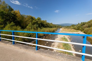 Güneşli bir günde güzel bir dağ manzarası. Doğal kıyı şeridi. Nehirde az miktarda su var. Beskid Dağları. Mavi ve beyaz balustrades 'li köprü. Sola Nehri. 