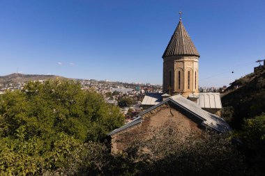 Bir tepenin üstünde Tiflis 'in çarpıcı manzarasını sunan tarihi bir kilise duruyor. Açık mavi gökyüzü ve arka plandaki şehir manzarası canlı bir Gürcistan başkenti manzarası yaratıyor..