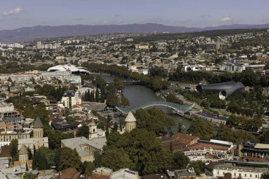 Tiflis 'e tepeden bakan canlı şehir, tarihi yapılarla nehir boyunca uzanan modern yapıları harmanlıyor. Manzara, berrak mavi gökyüzünün altında yemyeşil ve uzak tepeleri barındırır..