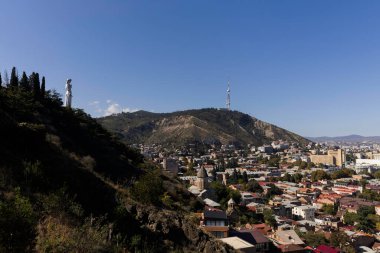 Ziyaretçiler, Tiflis 'in göz kamaştırıcı panoramik manzarasına hayran kalarak, aşağıdaki renkli şehri ve parlak mavi gökyüzünün altında göze çarpan heykel ile süslenmiş görkemli tepeleri yakalıyorlar..