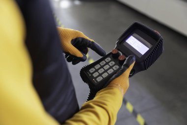 Güvenlik eldivenli bir işçi depodaki ekipmanları kontrol etmek için bir kontrol cihazı kullanıyor. Çevrede yerde endüstriyel elementler ve güvenlik işaretleri var.