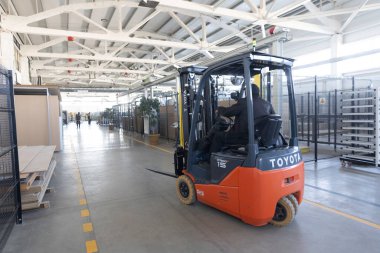 Düzenli malzeme ve ekipmanlarla dolu büyük bir depoda sorunsuzca hareket eden bir forklift görülüyor. İşçiler aktif olarak görevleriyle meşguller..