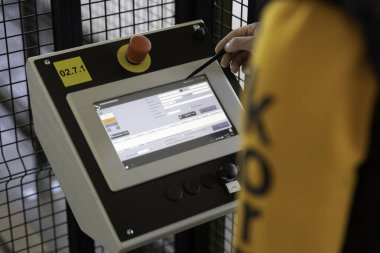 Bir operatör bir üretim tesisinde kontrol paneliyle karşılaşır. Ekran çeşitli ayarları yansıtırken, birey komut girmek için bir stil kullanır, kesin makine işlevini sağlar.