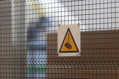 Parlak sarı bir uyarı levhası çite iliştirilmiş, inşaat alanına işaret ediyor. Arkaplan, kentsel alanda devam eden çalışmaları vurgulayarak, inşaat malzemelerinin bulanık ana hatlarını belirtiyor.