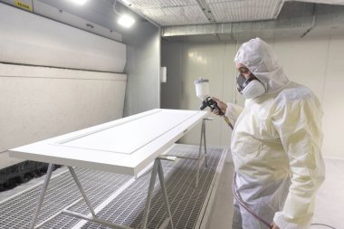 Tam koruma donanımlı bir işçi, temiz bir sprey boya kabinindeki beyaz bir kapıya dikkatlice sprey boya sıkar. Kontrollü ortam tehlikeleri en aza indirirken kalite sonuçlarını güvence altına alır.