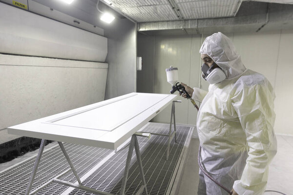 Работник в полном защитном снаряжении осторожно наносит краску на белую дверь в чистой распылительной кабинке. Контролируемая среда обеспечивает качественные результаты при минимизации рисков.