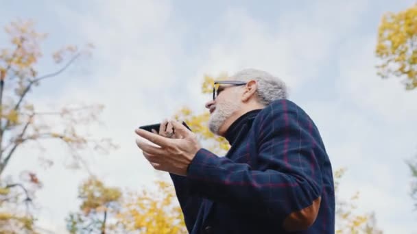 白髪と髭を生やした白人の中年男性が眼鏡をかけ 公園に立って電話で話している正式な服を着ていた カメラの回転 水平屋外ビデオ 高品質4K映像 — ストック動画