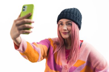 Makyajlı genç pembe saçlı kız gülümsüyor ve akıllı telefonuyla selfie çekiyor. Orta ölçekli yakın çekim stüdyosu beyaz arka plan çekiyor. Yüksek kalite fotoğraf
