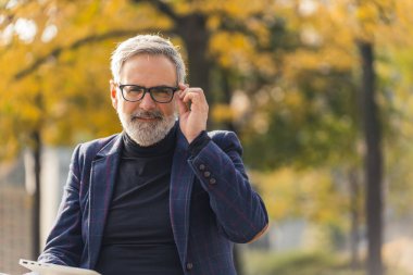 Modern ve kendine güvenen sakallı, gri saçlı, takım elbiseli, gözlüklü, güz yapraklarıyla parkta oturan, dizüstü bilgisayarı üzerinde çalışan yetişkin bir girişimci. Yüksek kalite fotoğraf