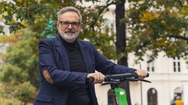 Milenyum çağının modern sakallı, gri saçlı, gözlüklü olgun bir adam, park alanında elektro scooterıyla dikiliyor. Yüksek kalite fotoğraf