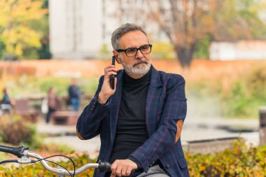 Sakallı, gri saçlı, olgun bir adam işe giderken parkta bisiklet sürerken telefonla birini arıyor. Yüksek kalite fotoğraf