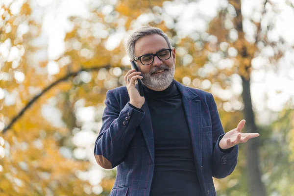 Stylish handsome bearded grey-haired mature man in a suit speaking with his friend on the phone. Fall foliage in the park. High quality photo