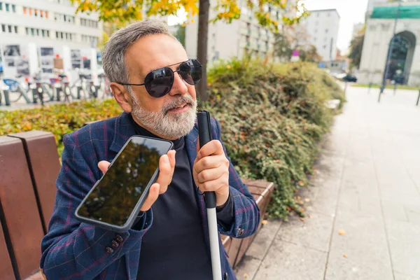 Elegant blind bearded grey-haired mature man with dark sunglasses on holding a walking stick, enjoying fall season in the park and talking with somebody through a speakerphone. High quality photo