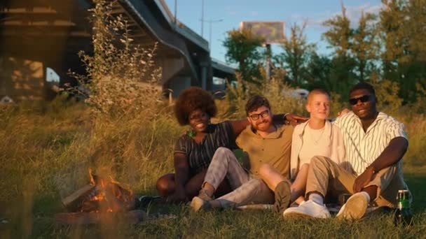 由四个不同种族的人组成的友好小组坐在一座城市桥下的草地上 享受篝火的温暖 听音乐 喝啤酒 高质量的4K镜头 — 图库视频影像