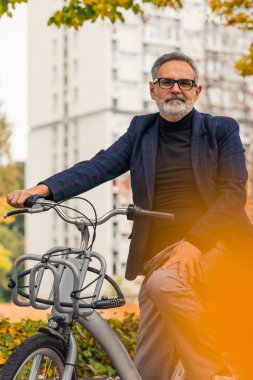 Beyaz saçlı, orta yaşlı, sakallı, gözlüklü, şık elbiseli, kiralık bisikletli bir adam parkta duruyor. İşe bisikletle gidiyorum. Dikey açık çekim. Yüksek kalite fotoğraf