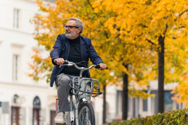 Gri saçlı, gözlüklü ve takım elbiseli orta yaşlı bir adam arka planda sonbahar ağaçlarıyla işe bisikletle gidiyor. Sağlıklı şehir yaşam tarzı. Yatay açık hava çekimi. Yüksek kalite fotoğraf