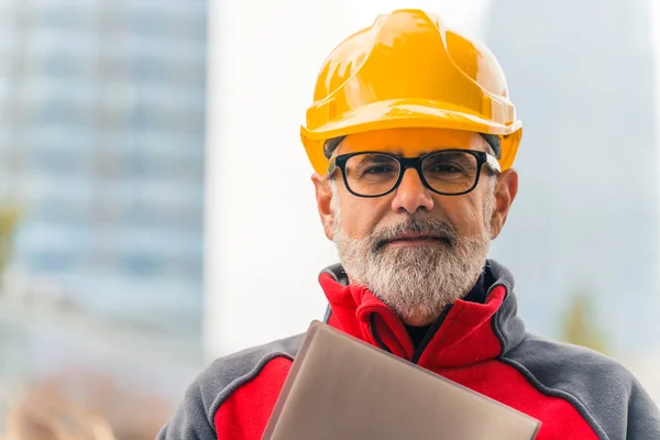 Middle-aged construction site manager wearing glasses and hard hat holding documents looking confidently into camera. Horizontal outdoor shot. High quality photo