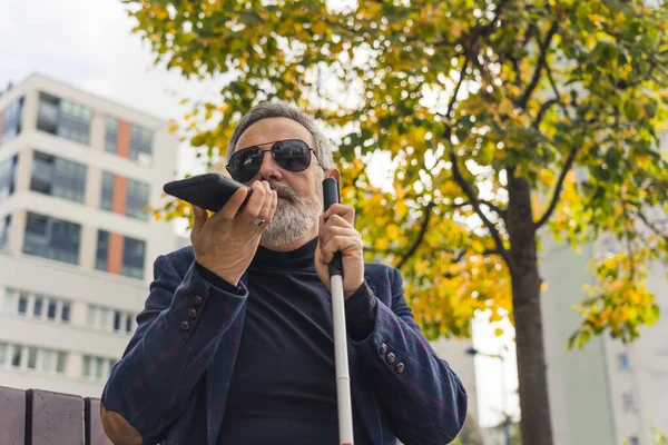 Middle-aged blind man with grey hair and beard wearing sunglasses and suit sitting on bench holding white cane and talking into phone. Horizontal outdoor shot. High quality photo