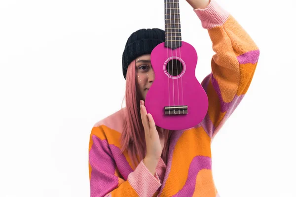 音乐激情的概念 中特写镜头 年轻的高加索粉红头发少女戴着黑色的便帽 穿着橙色和粉色的羊毛衫 手持新的炽热的粉色羽衣 覆盖了她的一半 — 图库照片