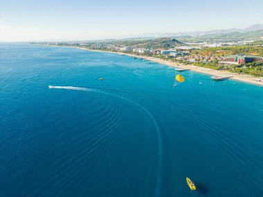 Okurcalar, Alanya, Türkiye. Geniş mavi Akdeniz manzaralı jet ski 'ler ve kıyıya yakın tekneler, lüks tatil köylerinin güneşli kumlu plajları, arka planda dağlar. Yüksek kalite fotoğraf