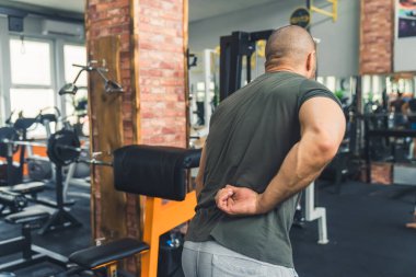 Arkaya bakan, kimliği belirsiz, beyaz tenli, siyah tişörtlü, profesyonel spor aletlerinin yanında duran ve kol kaslarını sıkılaştıran kel bir erkek. Spor salonunun içi. Yüksek kalite fotoğraf