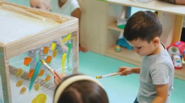 Anaokulu öğrencileri çocuk odasında eğleniyor ve fırçalarla resim yapıyorlar, raflar oyuncaklarla dolu. Yüksek kalite 4k görüntü